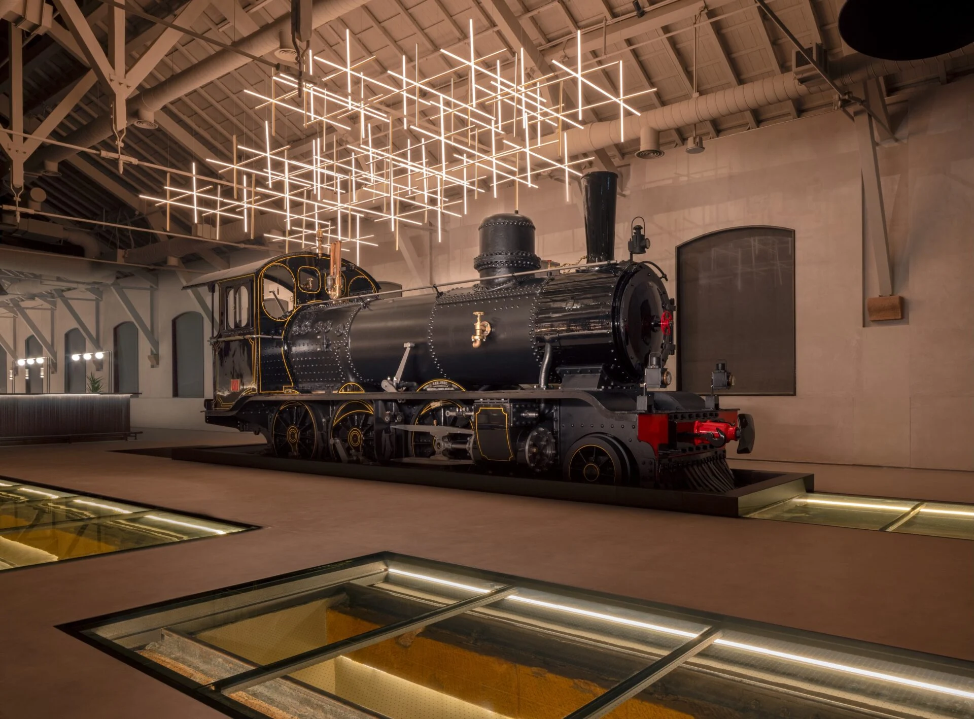Storica locomotiva a vapore nera esposta al chiuso sotto un'installazione luminosa a griglia e pannelli di pavimento in vetro – The Chedi Hegra, AlUla, Arabia Saudita, 2024 – architettura alberghiera