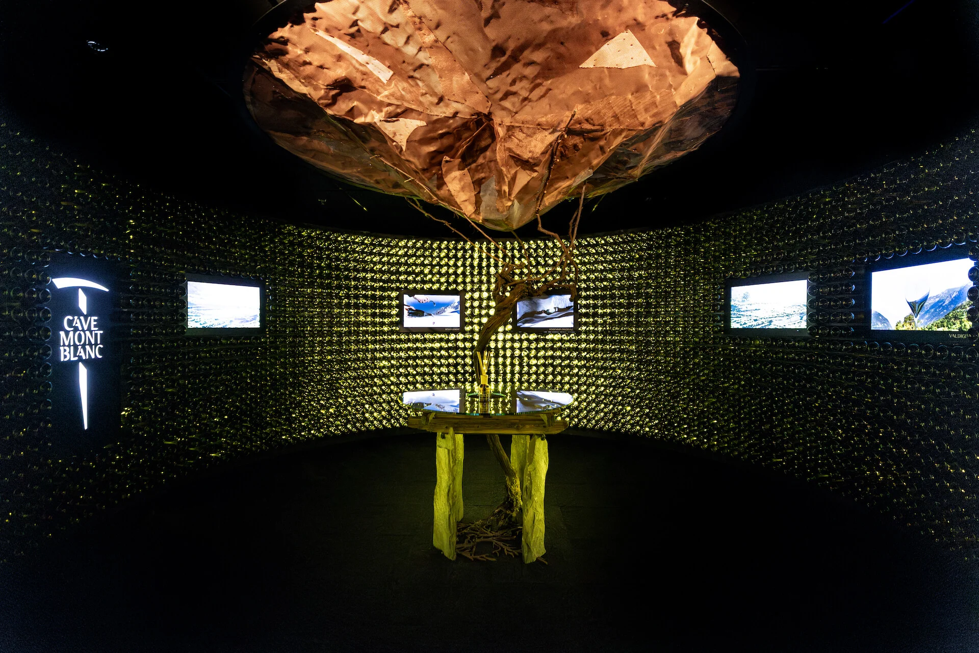 Scultura sospesa color rame sopra il tavolo con la vite, con schermi video inseriti nelle pareti di bottiglie – Cave Mont Blanc allo Skyway, Courmayeur, Italia, 2025 – interior design e allestimento espositivo