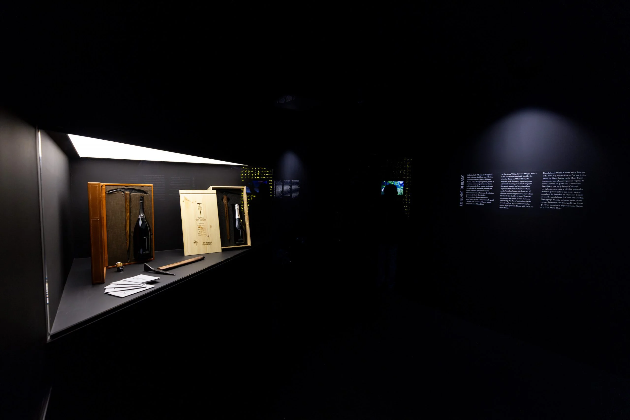 Vetrina in penombra con bottiglie in casse di legno e vecchi attrezzi, testi a parete che brillano nella sala buia – Cave Mont Blanc allo Skyway, Courmayeur, Italia, 2025 – interior design e allestimento espositivo
