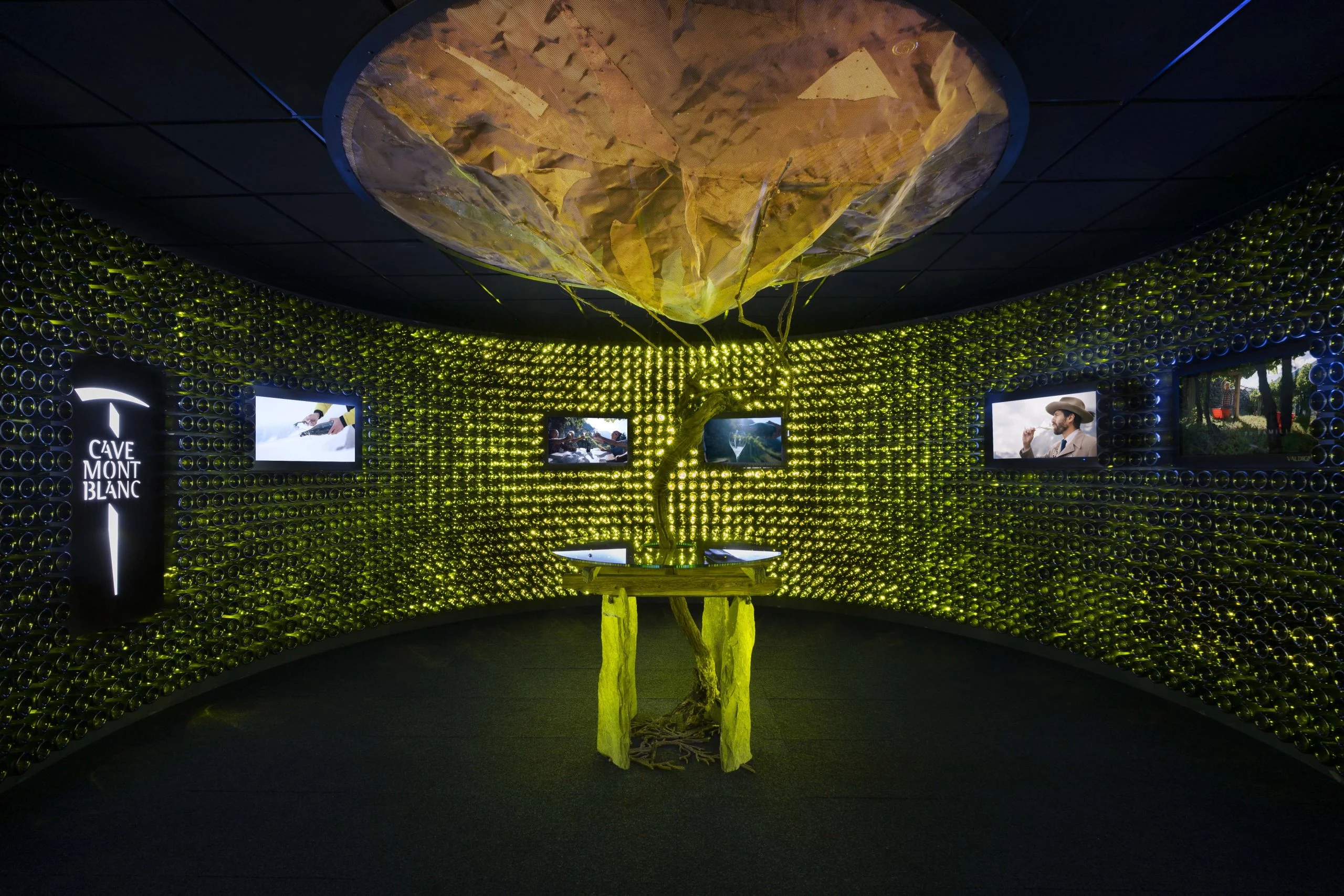 Sala circolare rivestita di bottiglie verdi retroilluminate, una vite su un tavolo sotto una scultura sospesa simile a una roccia – Cave Mont Blanc allo Skyway, Courmayeur, Italia, 2025 – interior design e allestimento espositivo