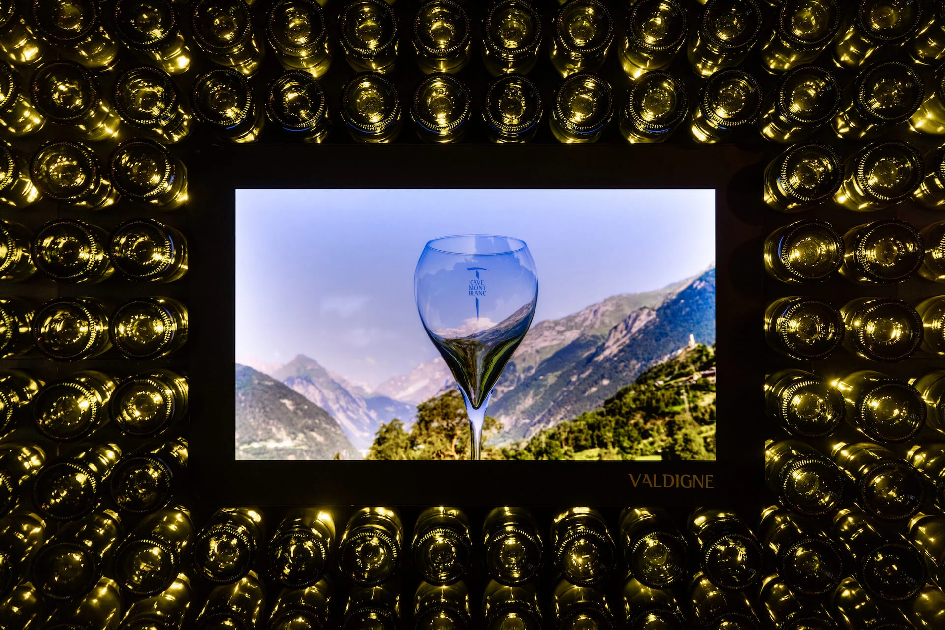 Dettaglio di uno schermo incorniciato da fondi di bottiglia, con un calice di vino contro una valle montana – Cave Mont Blanc allo Skyway, Courmayeur, Italia, 2025 – interior design e allestimento espositivo