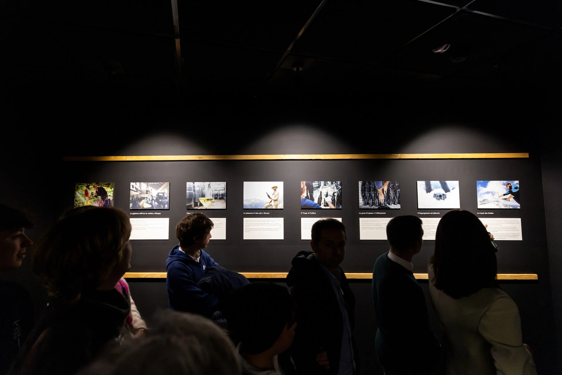 Visitatori davanti a una fila di fotografie retroilluminate con didascalie su una parete scura – Cave Mont Blanc allo Skyway, Courmayeur, Italia, 2025 – interior design e allestimento espositivo