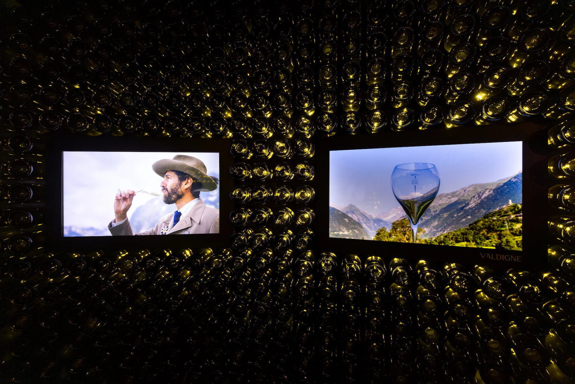 Due schermi inseriti nella parete di bottiglie: un uomo che degusta il vino e un calice contro le vette alpine – Cave Mont Blanc allo Skyway, Courmayeur, Italia, 2025 – interior design e allestimento espositivo