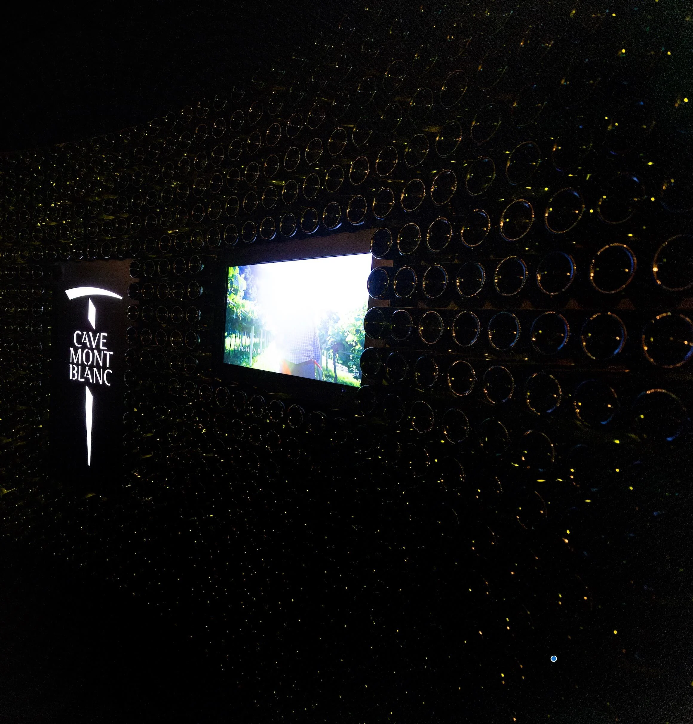 Parete scura di fondi di bottiglia con il logo illuminato Cave Mont Blanc e uno schermo con vigneti – Cave Mont Blanc allo Skyway, Courmayeur, Italia, 2025 – interior design e allestimento espositivo