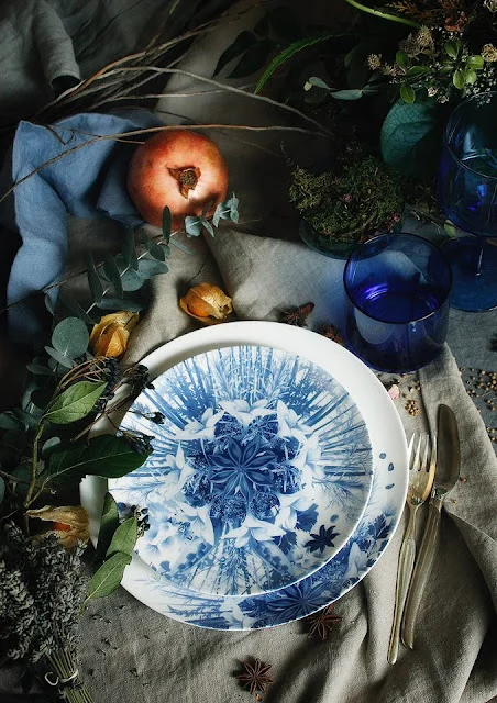 Vista dall'alto di piatti con decori blu, una melagrana, erbe aromatiche e un bicchiere blu su lino stropicciato – Servizio da tavola Botanica per Bitossi Home, 2017 – design di prodotto