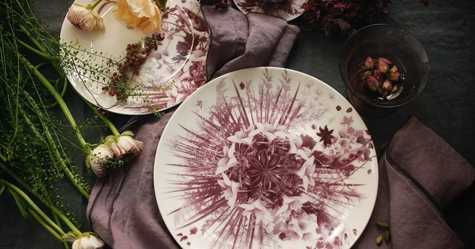 Vista dall'alto di un piatto con decoro botanico color prugna su lino grigio, con fiori e un bicchiere – Servizio da tavola Botanica per Bitossi Home, 2017 – design di prodotto