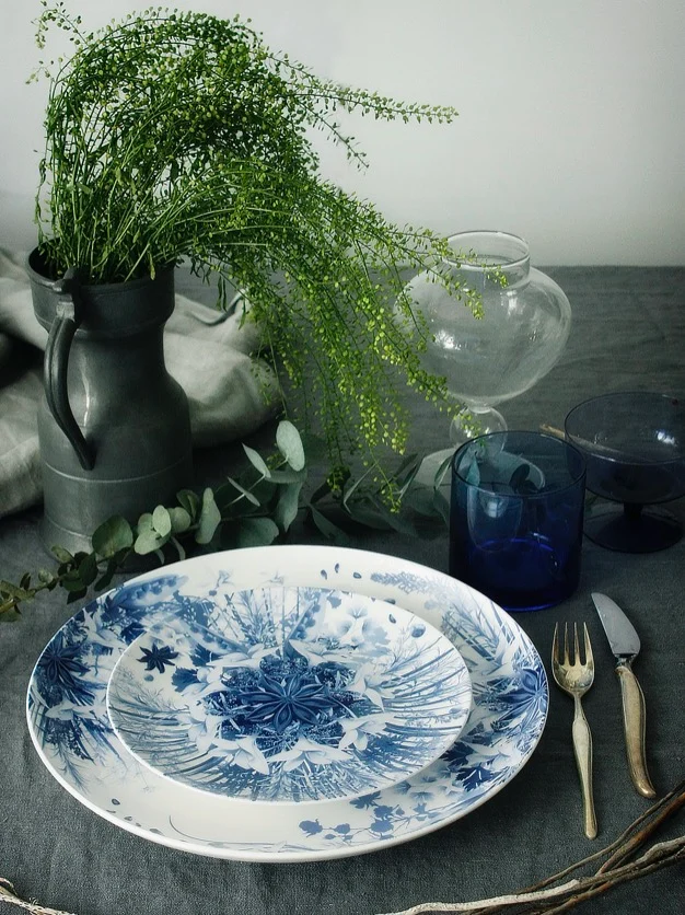 Piatti con decoro botanico blu impilati su una tovaglia grigia con posate, bicchieri blu e una brocca di fronde verdi – Servizio da tavola Botanica per Bitossi Home, 2017 – design di prodotto