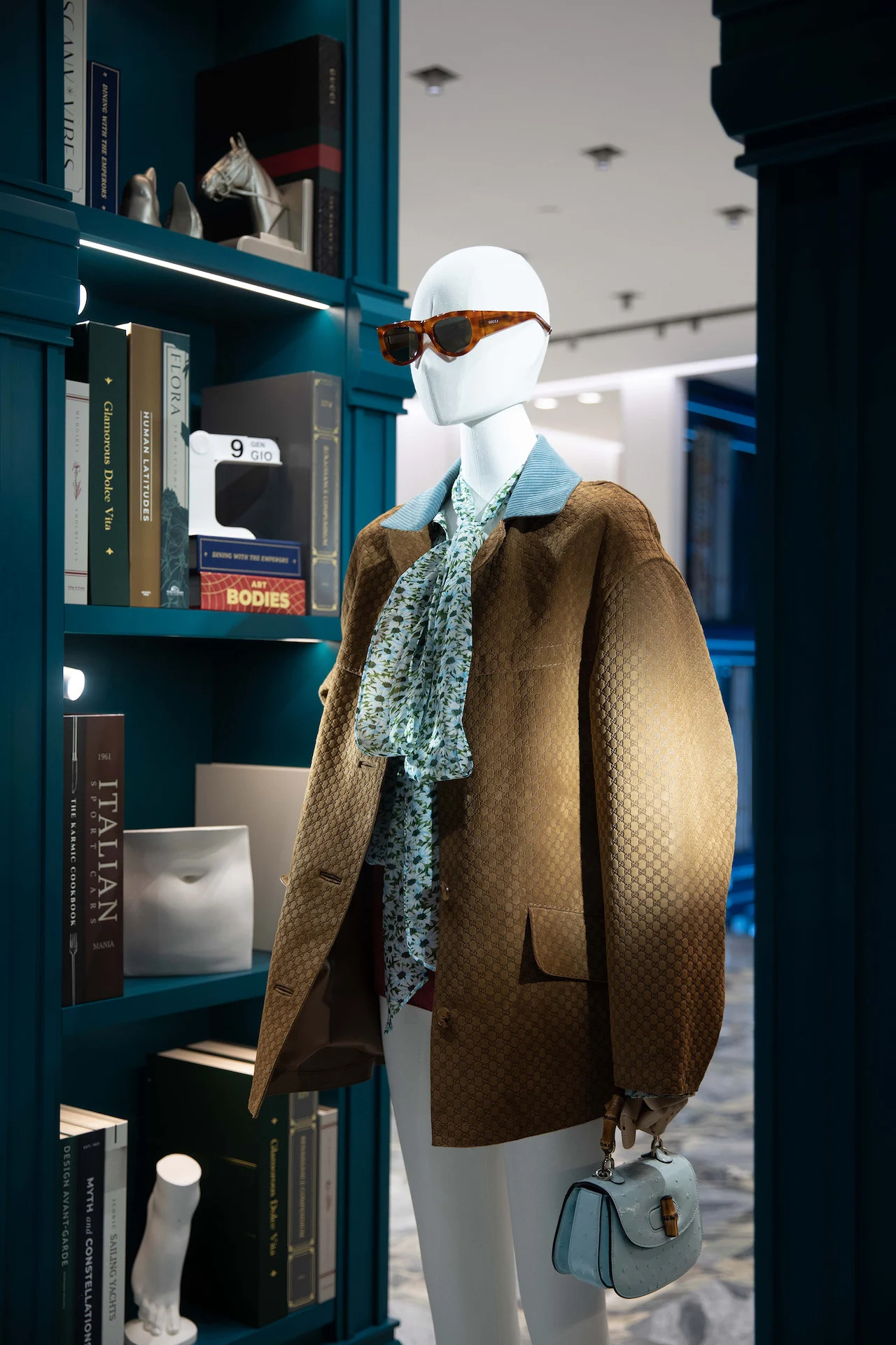Manichino con giacca in camoscio e occhiali da sole accanto a una libreria verde-azzurro con libri d'arte e oggetti – Vetrine Gucci Endless Narratives, 2025 – retail design