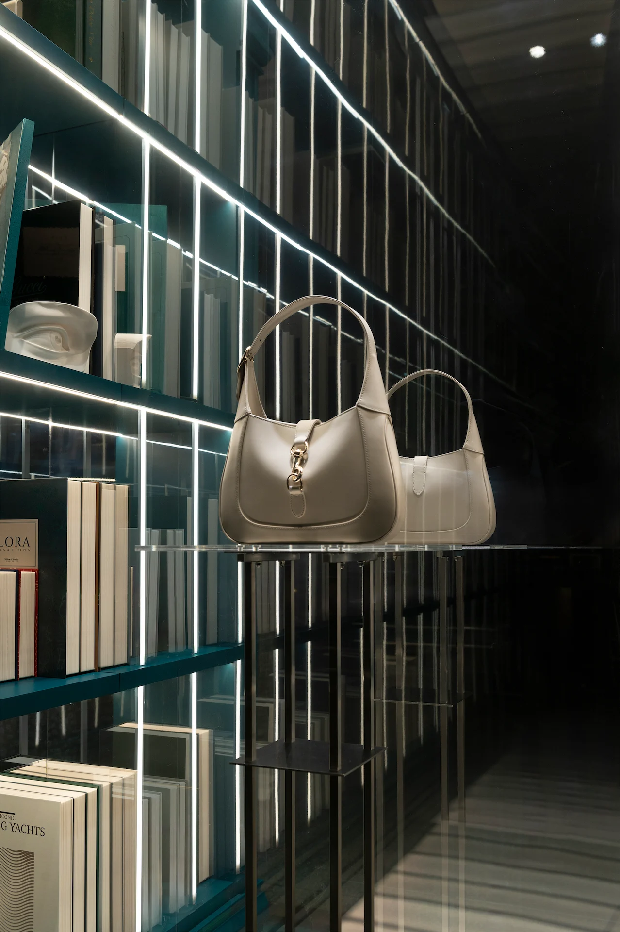 Due borse color crema su un ripiano di vetro contro librerie a specchio con strisce di luce – Vetrine Gucci Endless Narratives, 2025 – retail design