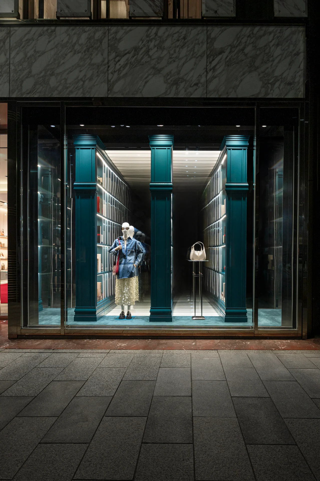 Vista notturna di una vetrina Gucci con un manichino e una borsa bianca tra colonne e librerie verde acqua – Vetrine Gucci Endless Narratives, 2025 – retail design