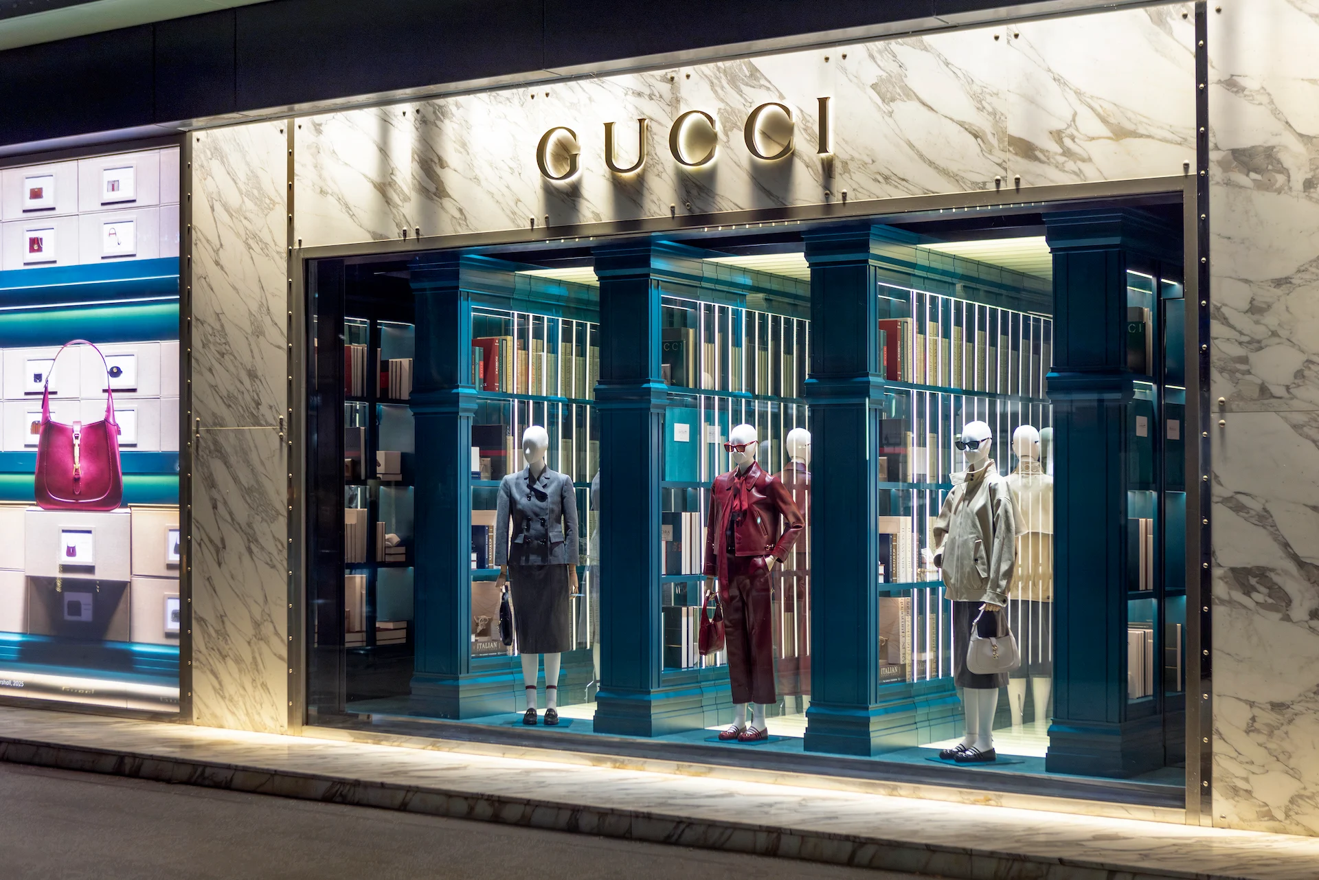 Vista di scorcio di una vetrina Gucci con tre manichini e librerie verde acqua illuminate sotto un logo dorato – Vetrine Gucci Endless Narratives, 2025 – retail design
