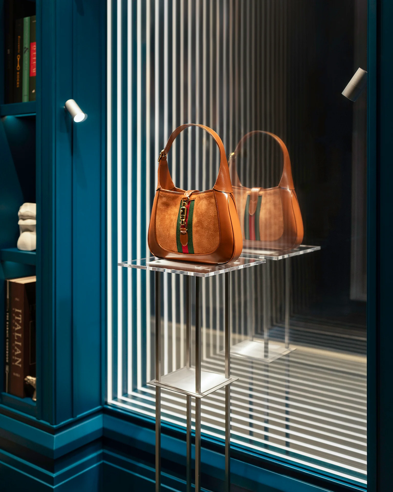 Dettaglio di una borsa Gucci in camoscio color cuoio su un supporto in acrilico, riflessa in uno specchio con strisce di luce verticali – Vetrine Gucci Endless Narratives, 2025 – retail design