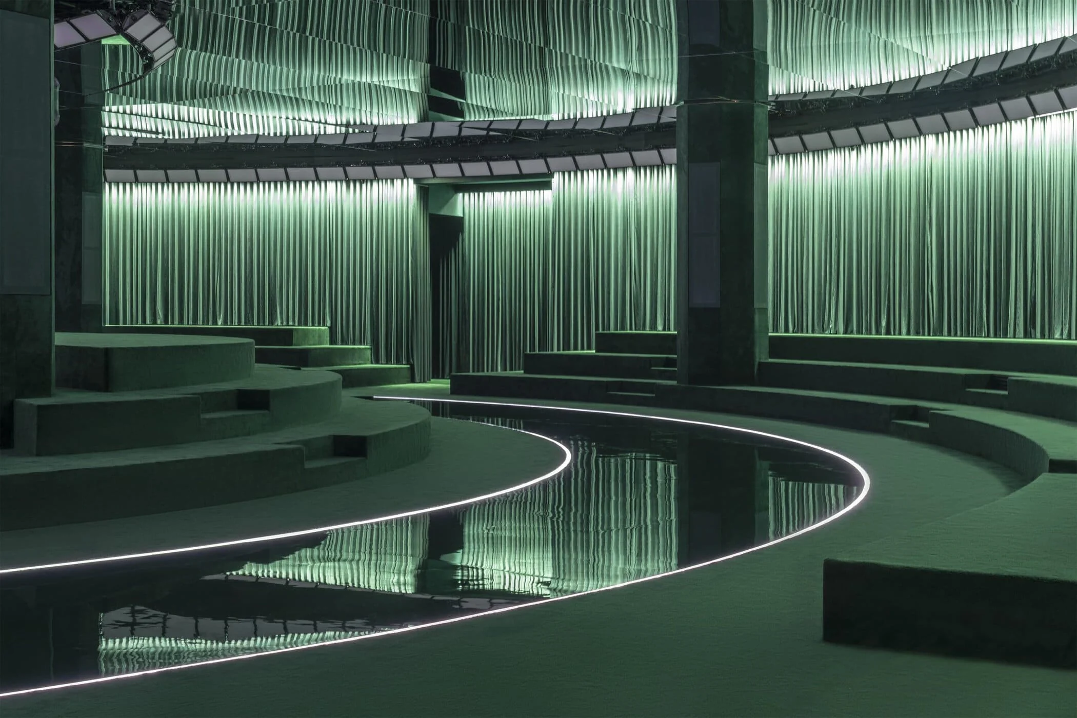 Passerella curva in specchio nero bordata di luce, che si snoda tra le gradinate in velluto verde – Gucci Autunno/Inverno 2025, Milano, Italia – scenografia per sfilate