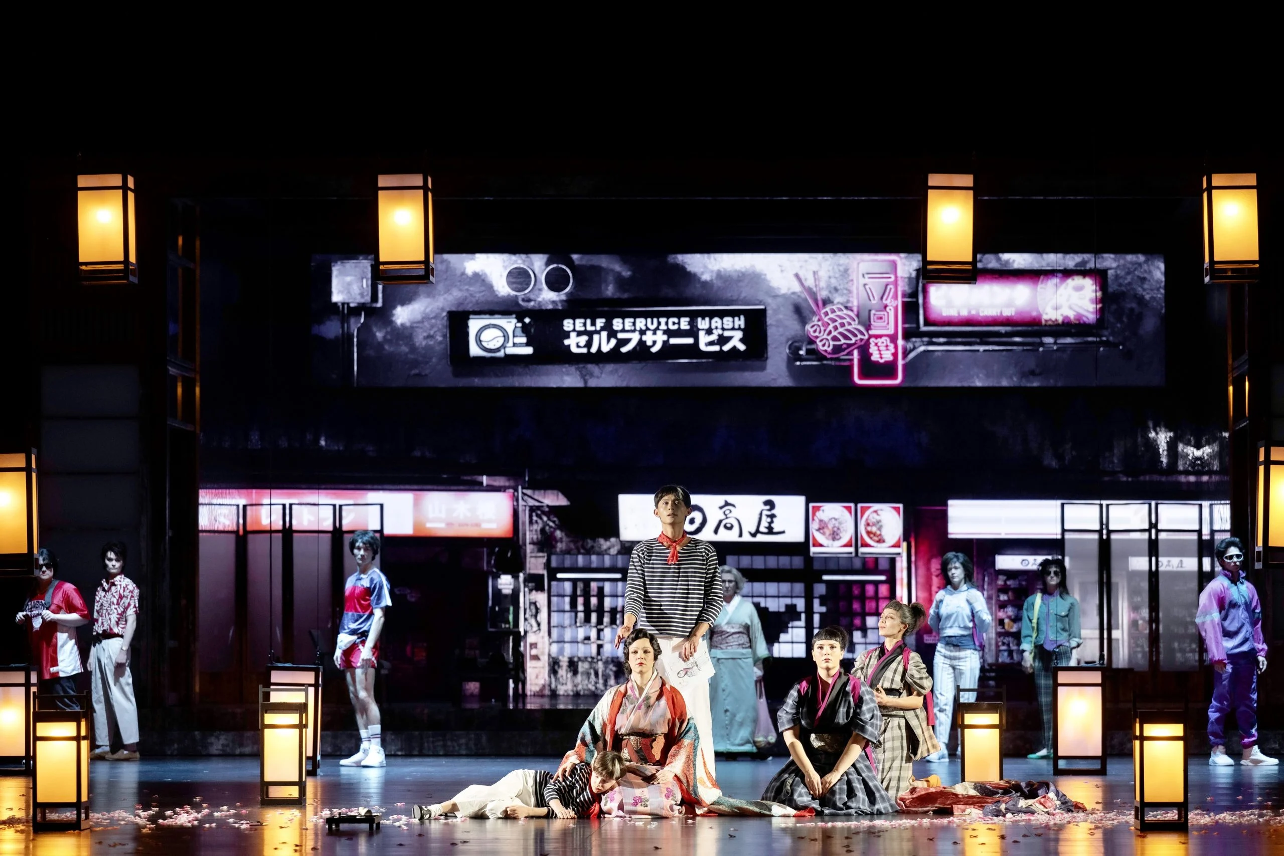 Coro e solisti nella scenografia della strada giapponese moderna, illuminati da lanterne di carta lungo il bordo del palco – Madama Butterfly al Festspielhaus di Baden-Baden, Germania, 2025 – scenografia d'opera