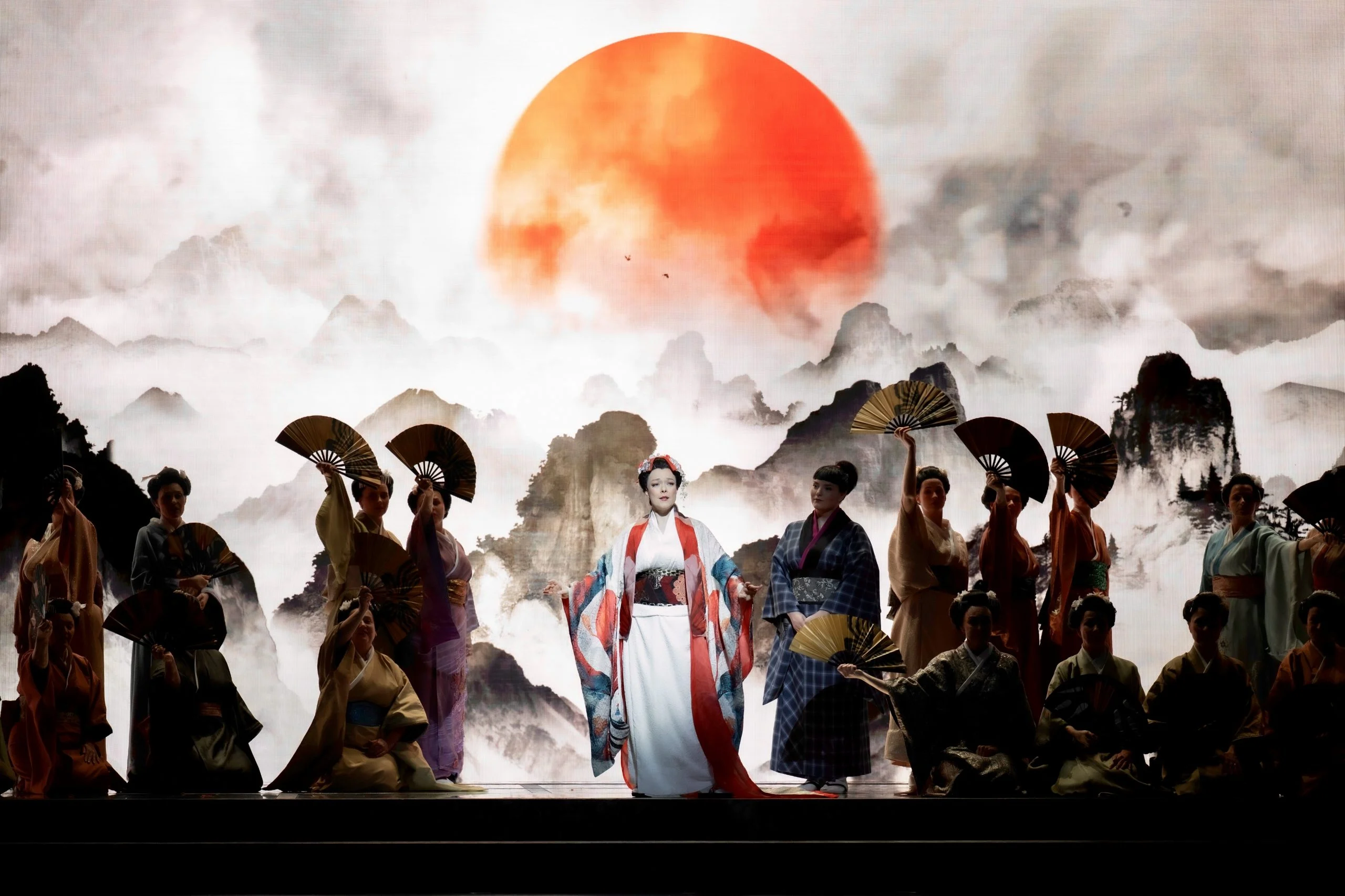 Soprano in kimono bianco e rosso tra donne con ventagli, davanti alla proiezione di un sole rosso su montagne nebbiose – Madama Butterfly al Festspielhaus di Baden-Baden, Germania, 2025 – scenografia d'opera