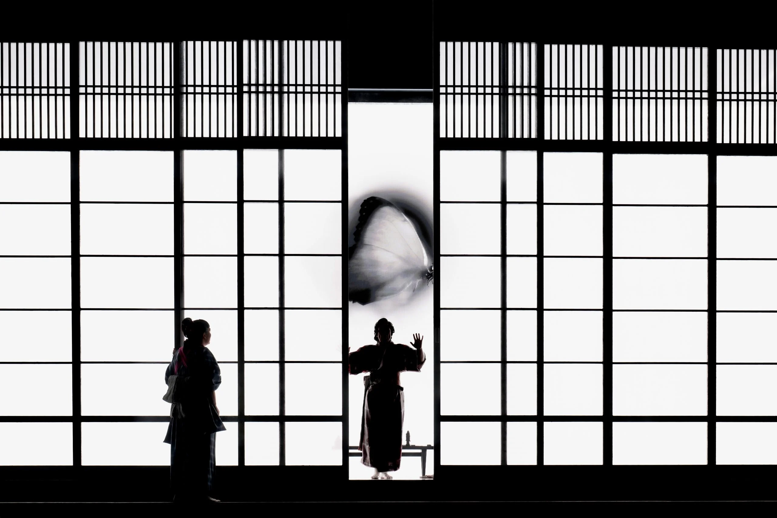 Silhouette di due donne davanti a shoji retroilluminati, una farfalla gigante proiettata sul pannello centrale – Madama Butterfly al Festspielhaus di Baden-Baden, Germania, 2025 – scenografia d'opera