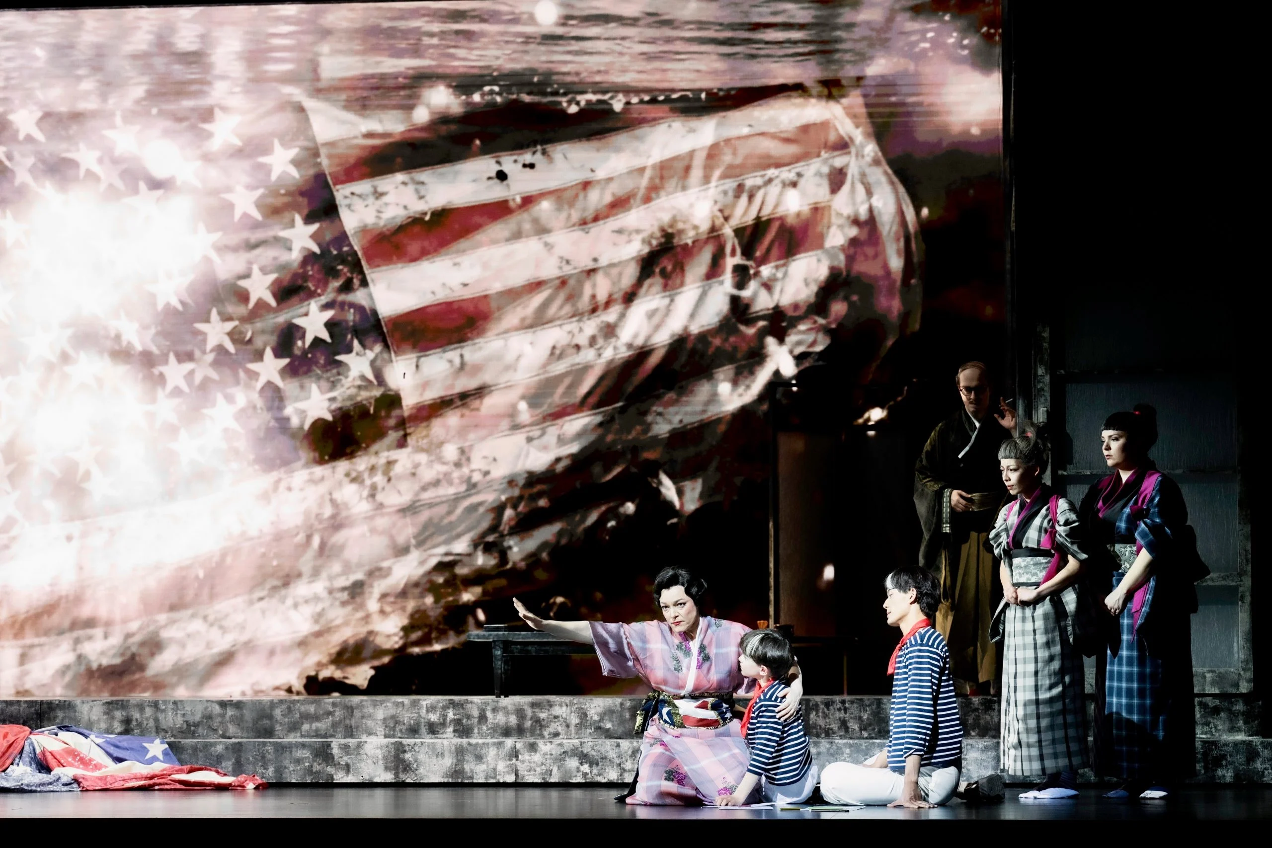 Cantanti in kimono sul palco davanti a una gigantesca bandiera americana proiettata, una bandiera accartocciata a terra – Madama Butterfly al Festspielhaus di Baden-Baden, Germania, 2025 – scenografia d'opera
