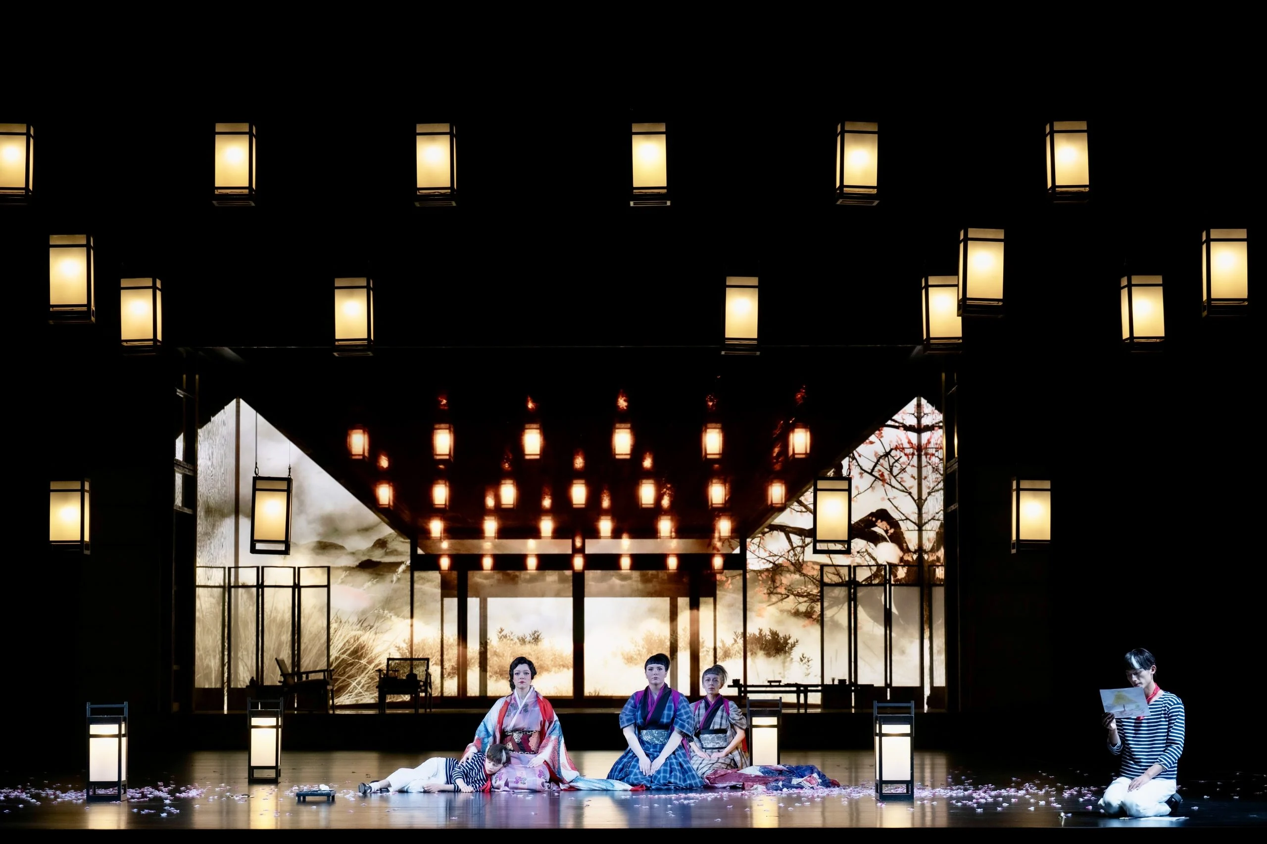 Tre donne inginocchiate su un palco cosparso di petali, incorniciate da una griglia di lanterne appese e da un padiglione illuminato – Madama Butterfly al Festspielhaus di Baden-Baden, Germania, 2025 – scenografia d'opera