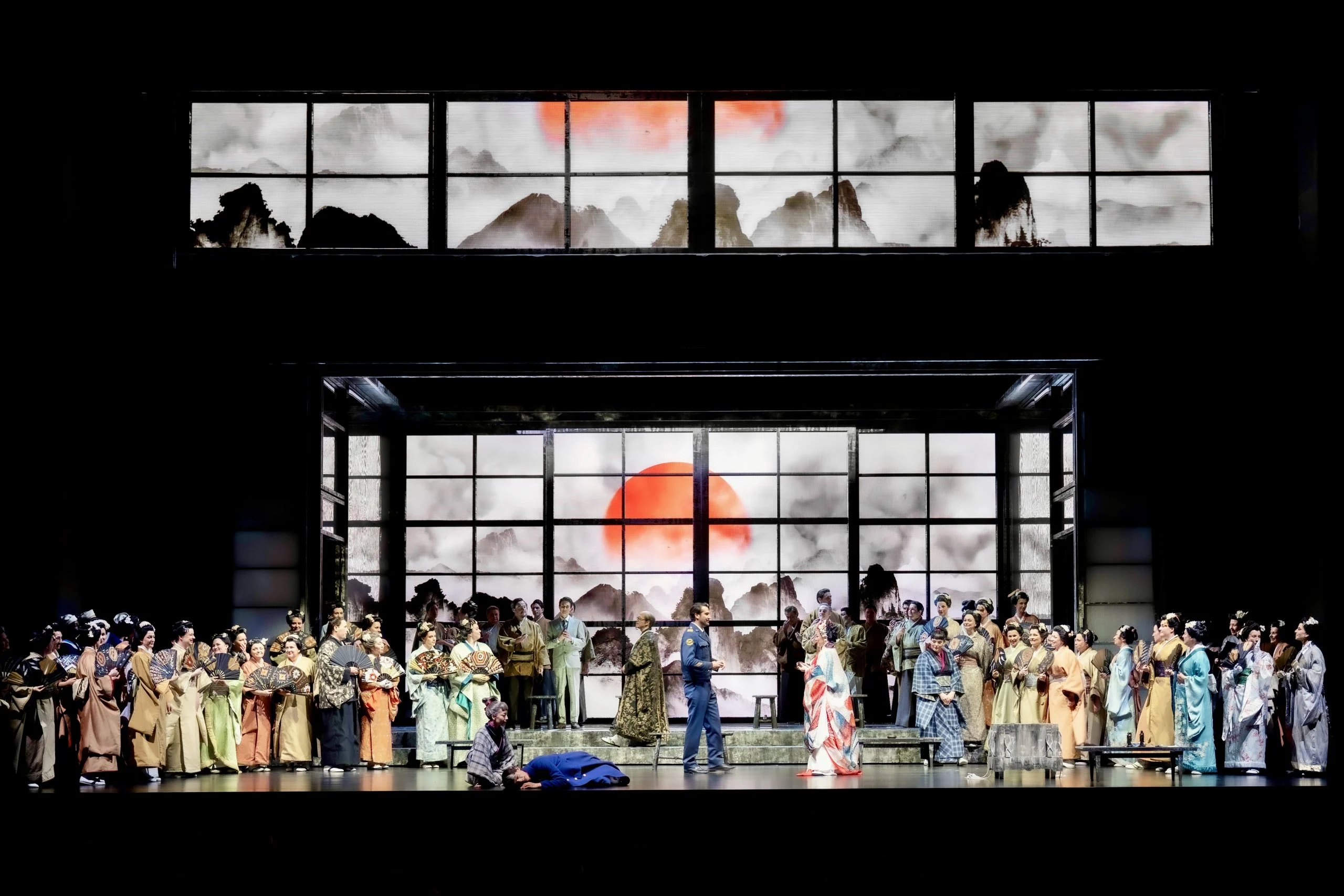 Il coro al completo in kimono sul palco davanti a schermi di vetro con un sole rosso su montagne a inchiostro – Madama Butterfly al Festspielhaus di Baden-Baden, Germania, 2025 – scenografia d'opera