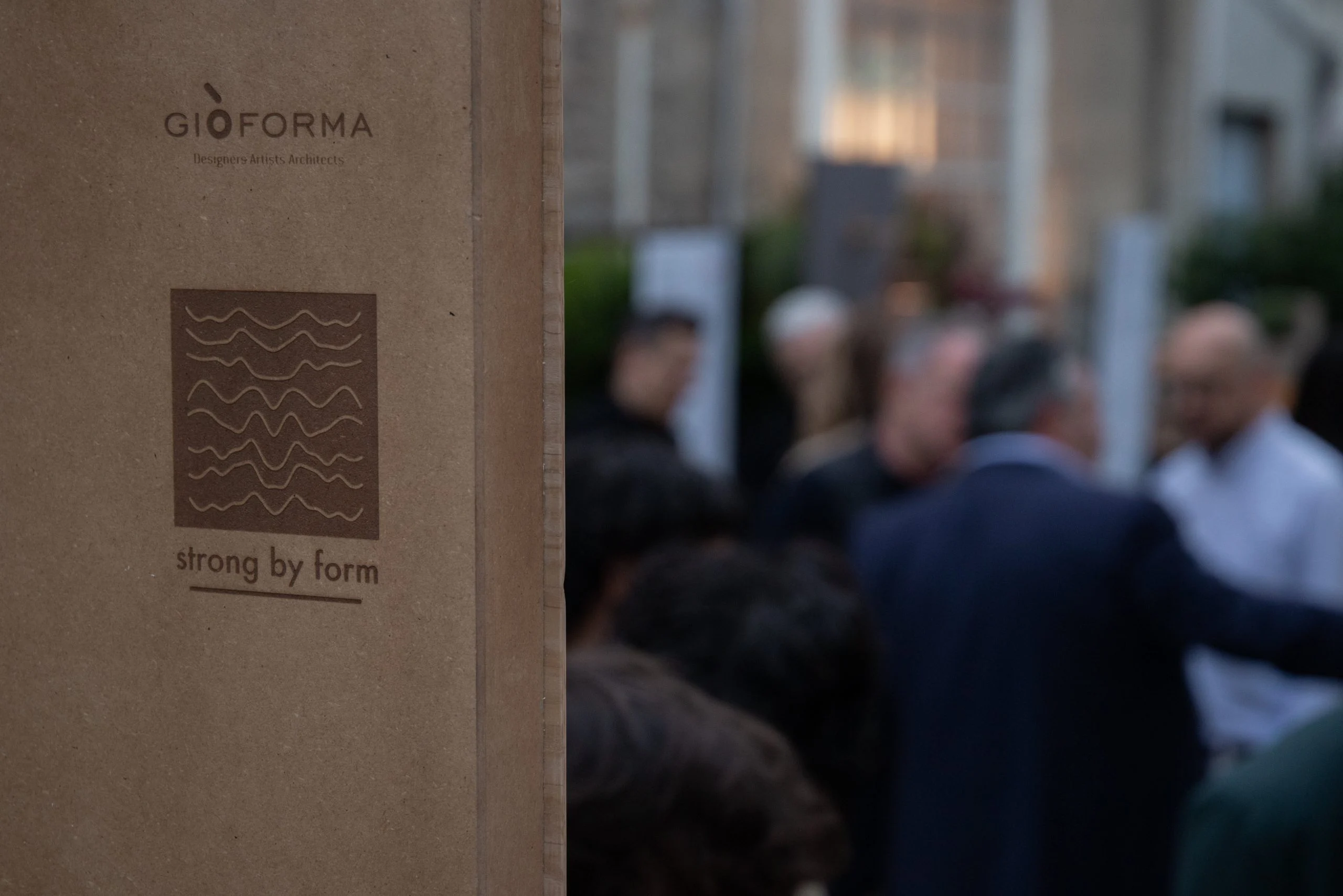 Scatola di cartone con il logo Strong by Form in primo piano, dietro ospiti che chiacchierano fuori fuoco – Everything Is a Party al Fuorisalone, Milano, Italia, 2025 – progettazione di eventi