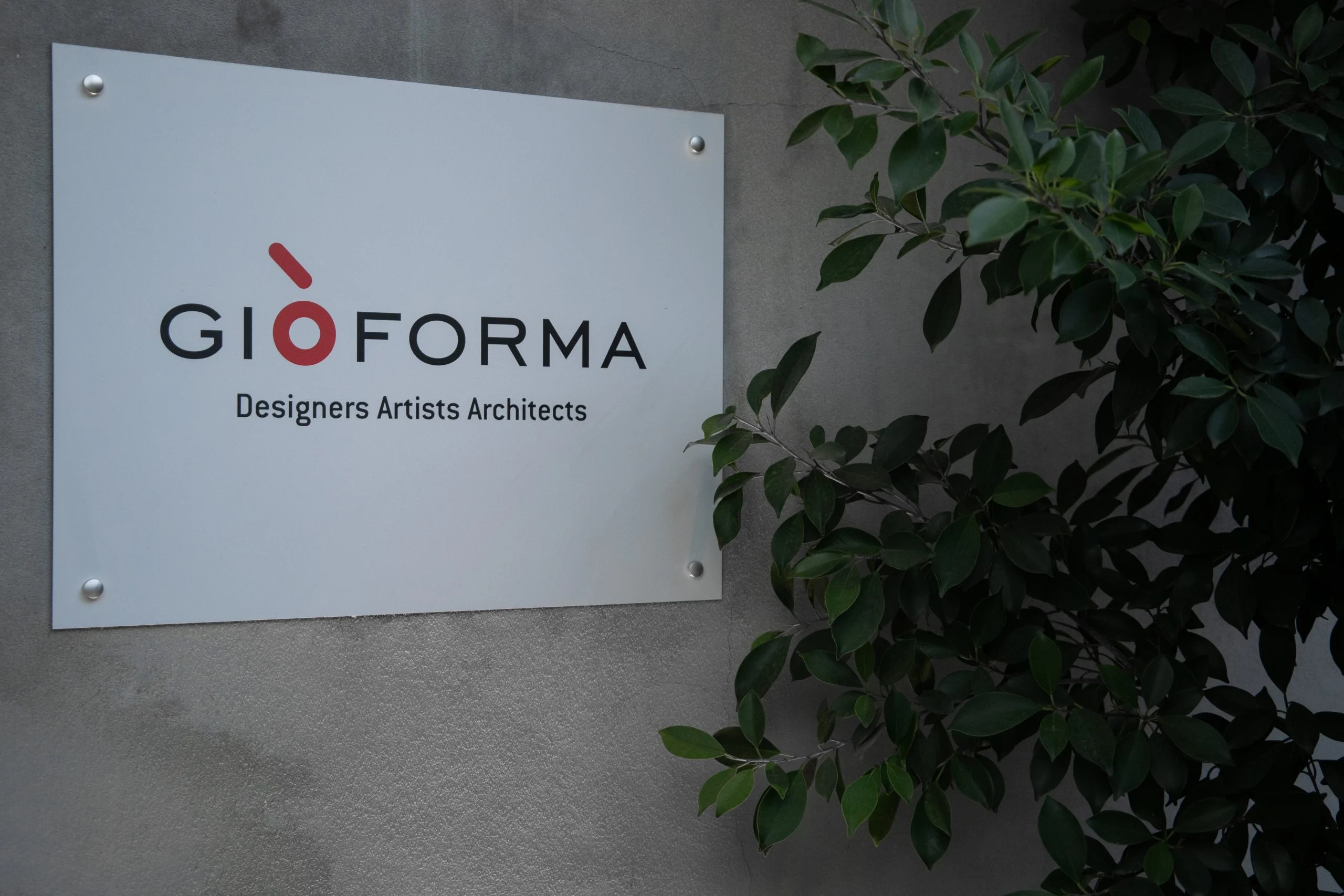 Insegna dello studio con logo e le parole Designers Artists Architects su una parete di cemento accanto a una pianta – Everything Is a Party al Fuorisalone, Milano, Italia, 2025 – progettazione di eventi
