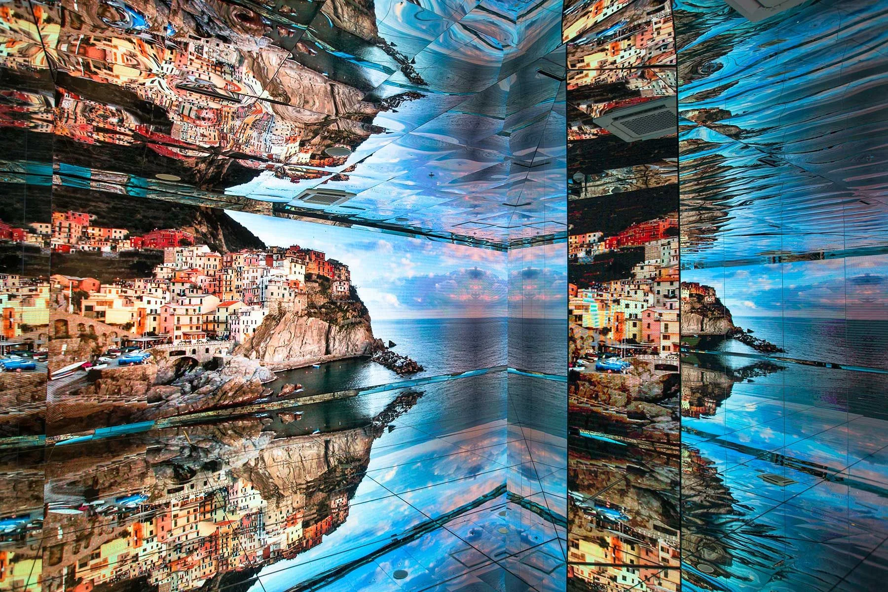 Sala a specchi con schermi che mostrano un colorato borgo marinaro a picco sulla scogliera, riflesso su ogni superficie – Padiglione Italia all'Expo 2015, Milano, Italia – allestimento espositivo