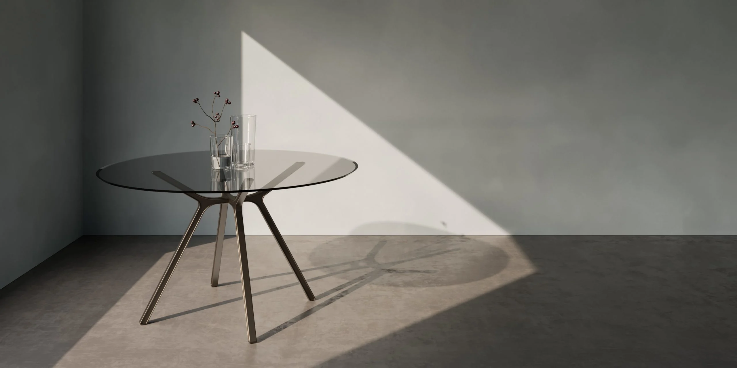 Tavolo rotondo in vetro con gambe metalliche inclinate in una stanza grigia attraversata da un raggio di sole – Collezione FRAME Tonino Lamborghini, Milano, Italia, 2025 – design di prodotto