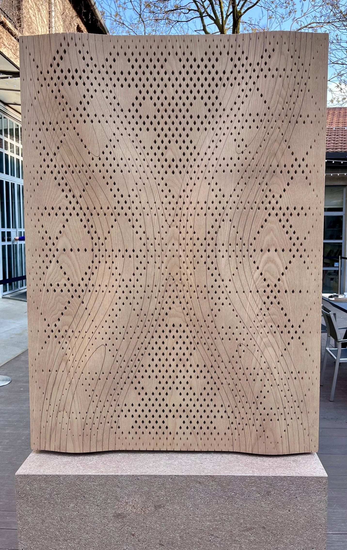 Vista frontale di un pannello acustico in legno ondulato e traforato su un basamento di pietra in un cortile – Pannelli Woodflow-Skin Strong by Form, Milano, Italia, 2025 – design di prodotto