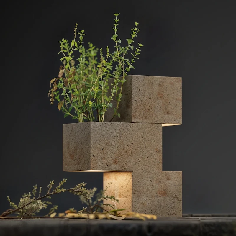 Lampada di tre blocchi di pietra sfalsati con una pianta che cresce in cima, luce che filtra dal basso – Arredi in porfido Porfido Pedretti, Milano, Italia, 2025 – design di prodotto