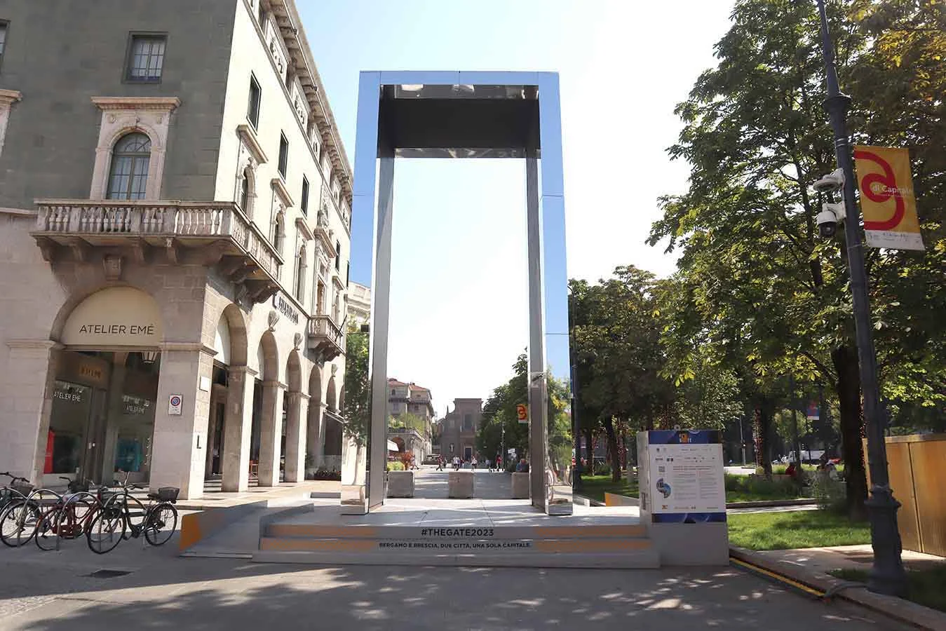 Portale THE GATE rivestito di specchi su una pedana in una piazza alberata in una giornata di sole – THE GATE 2023, Bergamo e Brescia, Italia – progettazione di installazioni
