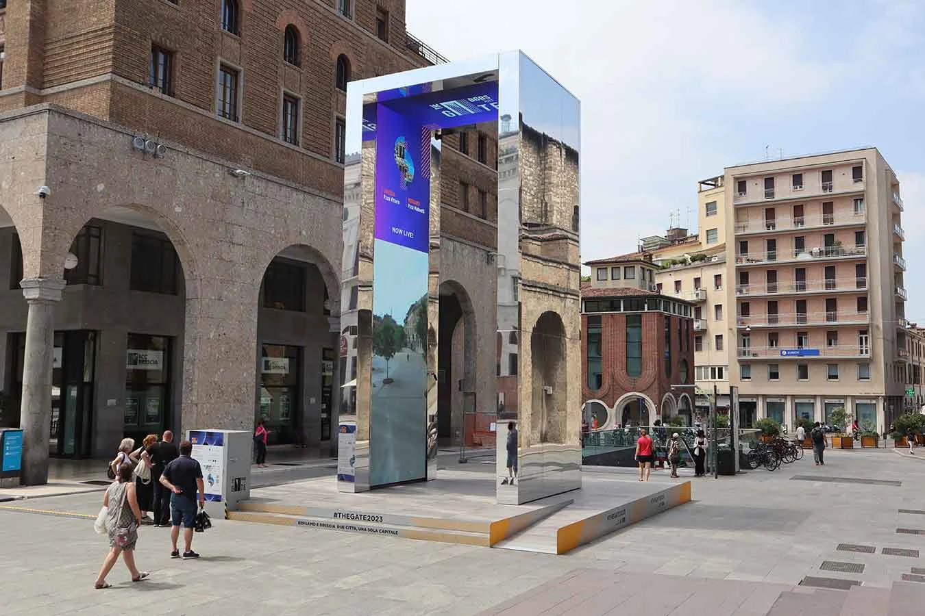 Portale rivestito di specchi che riflette edifici porticati, lo schermo LED interno con un video mentre passano persone – THE GATE 2023, Bergamo e Brescia, Italia – progettazione di installazioni