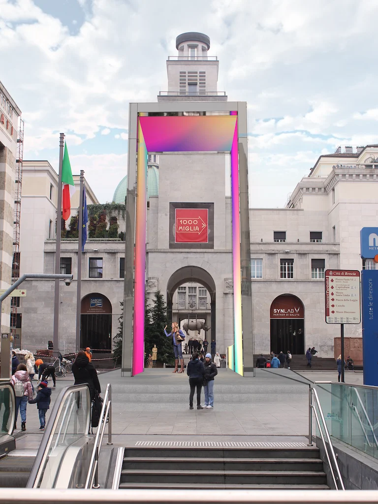 Portale THE GATE illuminato da schermi LED iridescenti in una piazza storica, con una torre dell'orologio inquadrata dietro – THE GATE 2023, Bergamo e Brescia, Italia – progettazione di installazioni