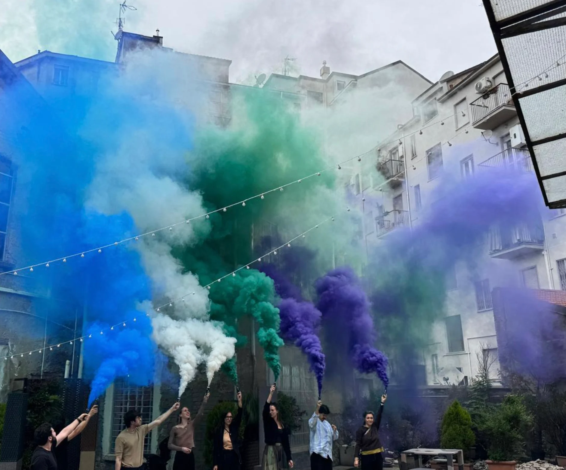 Persone con fumogeni blu, bianchi, verdi e viola in un cortile tra edifici cittadini – Cesare Cremonini Live25, Italia, 2025 – scenografia per concerti