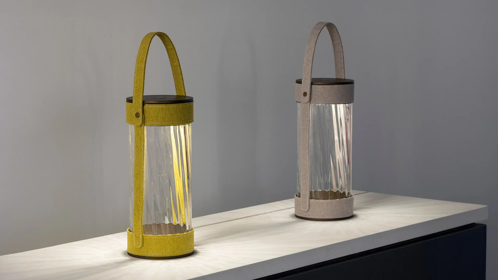 Due lanterne con cilindri in vetro rigato e cinghie di tessuto giallo e grigio su una mensola illuminata – Lanterna AlUla per Artemide, Milano, Italia, 2025 – lighting design