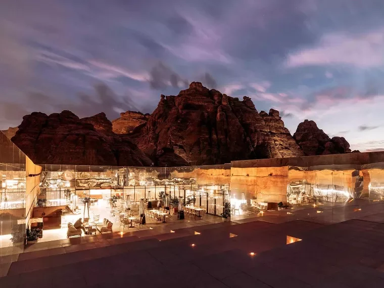 Terrazza con sedie in rattan e tavoli in marmo con lampade, parete vetrata e falesie riflesse alle spalle – Maraya Concert Hall, AlUla, Arabia Saudita, 2019 – architettura