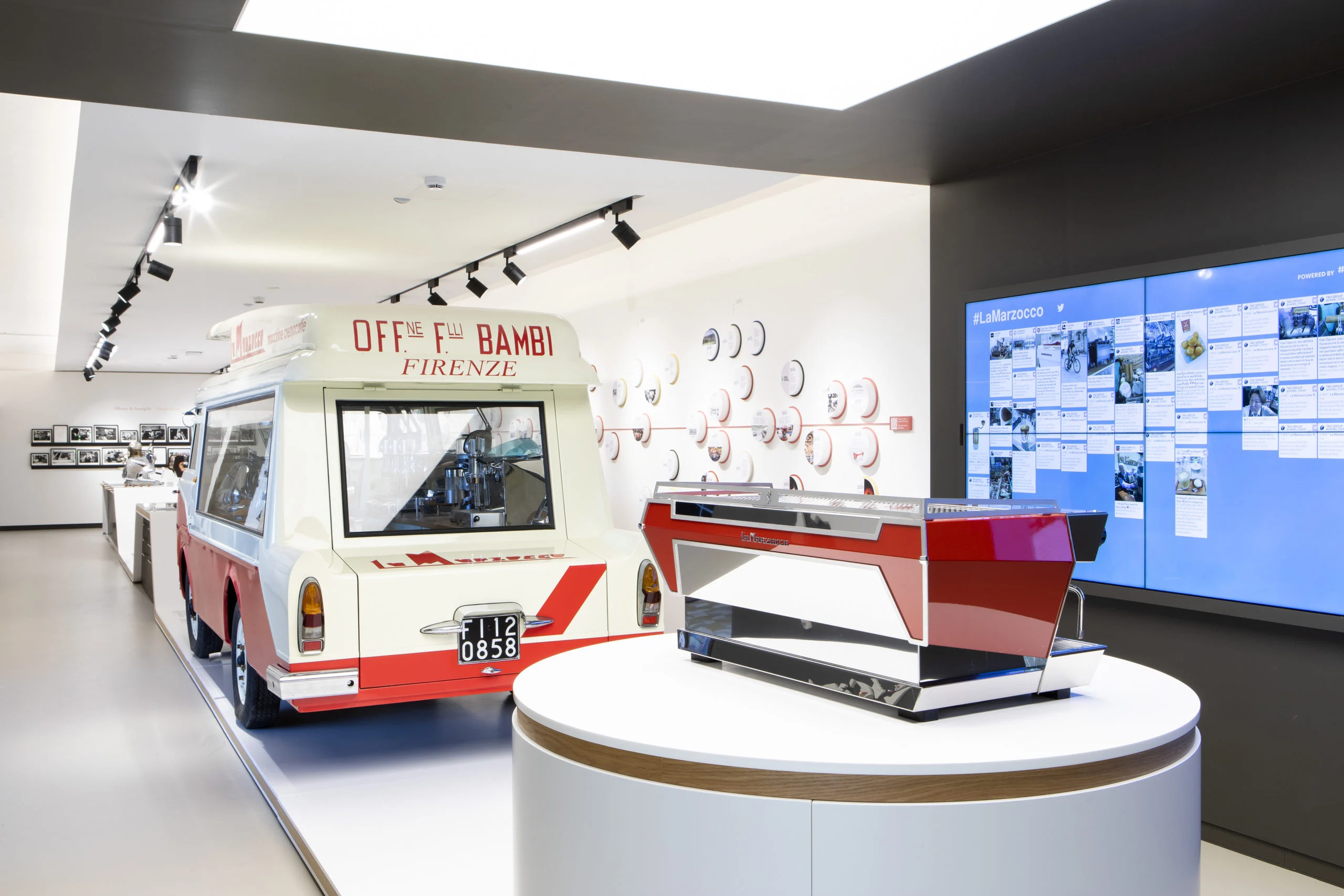 Macchina per espresso rossa su un basamento rotondo accanto a un furgone d'epoca e a una parete di foto digitali – La Marzocco Accademia del Caffè Espresso, Firenze, Italia, 2018 – interior design