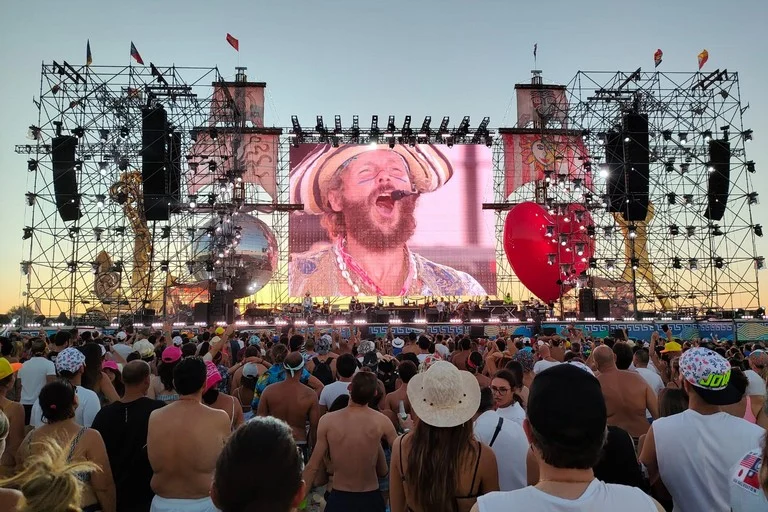Pubblico in spiaggia al tramonto davanti al palco, sullo schermo il cantante con un cappello di paglia, tra un cuore rosso e una palla a specchi – Jova Beach Party 2022, Italia – scenografia per concerti