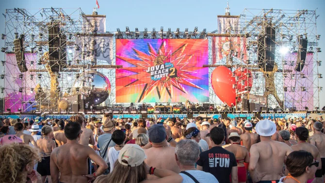 Pubblico in spiaggia di giorno davanti al palco del Jova Beach Party: torri di tubi, uno schermo con il logo colorato e un enorme cuore rosso – Jova Beach Party 2022, Italia – scenografia per concerti