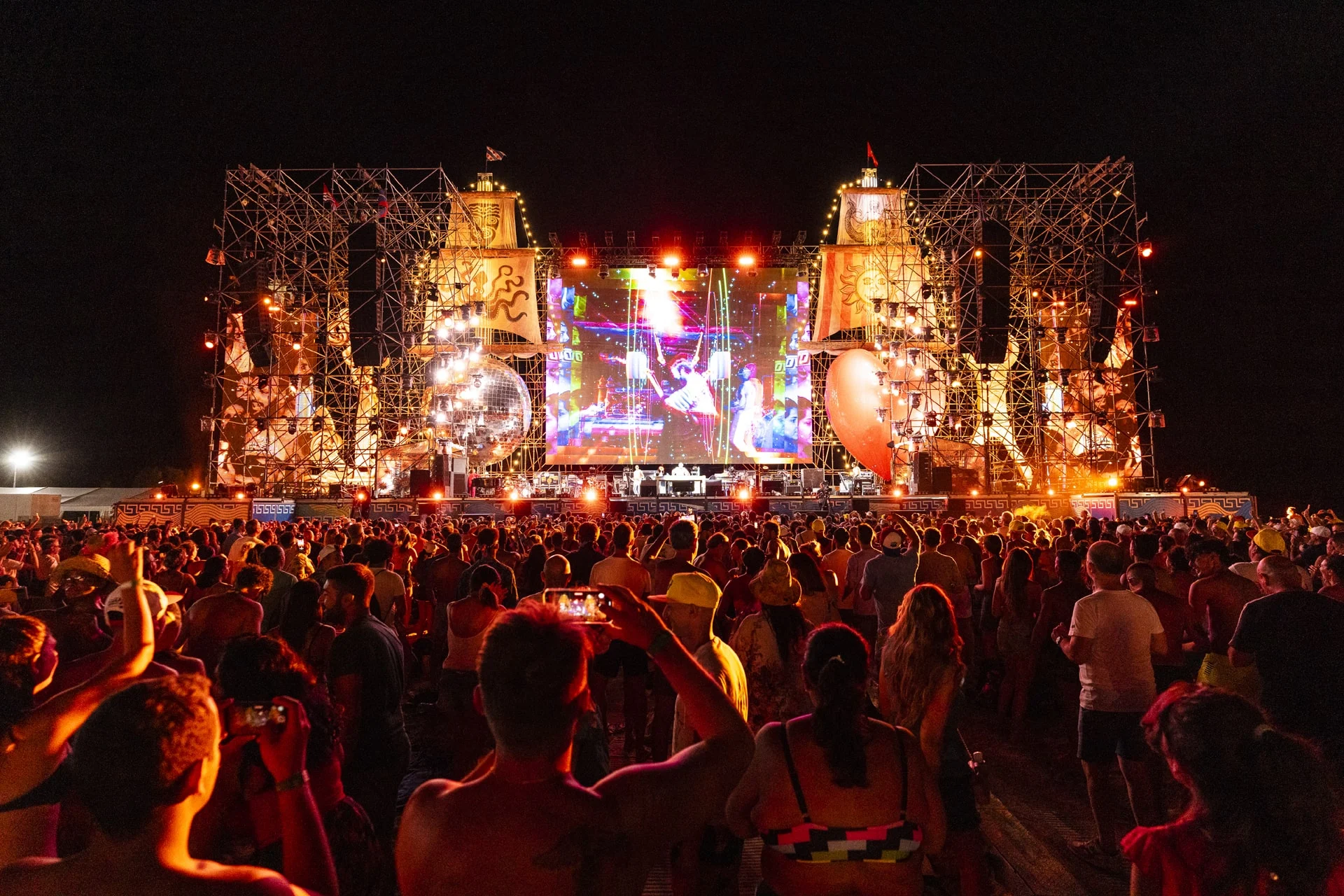 Vista notturna dal pubblico, telefoni alzati verso il palco di tubi illuminato con torri a forma di nave e uno schermo centrale – Jova Beach Party 2022, Italia – scenografia per concerti