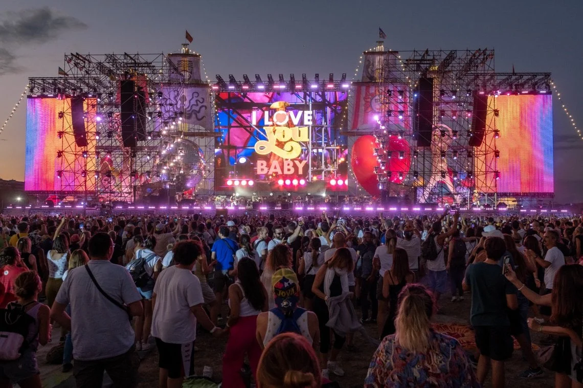 Pubblico al crepuscolo davanti al palco illuminato, con "I Love You Baby" sullo schermo centrale e schermi laterali colorati – Jova Beach Party 2022, Italia – scenografia per concerti