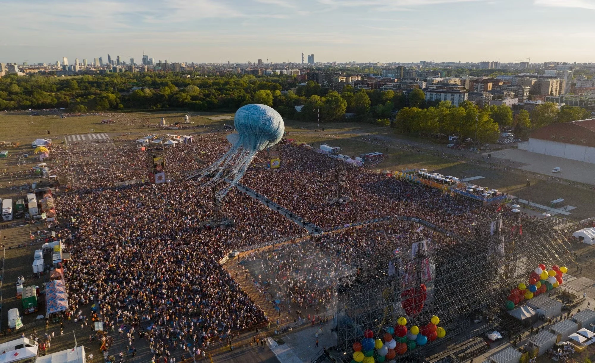 Vista aerea del pubblico del concerto in un parco, l'aerostato a medusa sospeso sopra e lo skyline della città sullo sfondo – Jova Beach Party 2022, Italia – scenografia per concerti