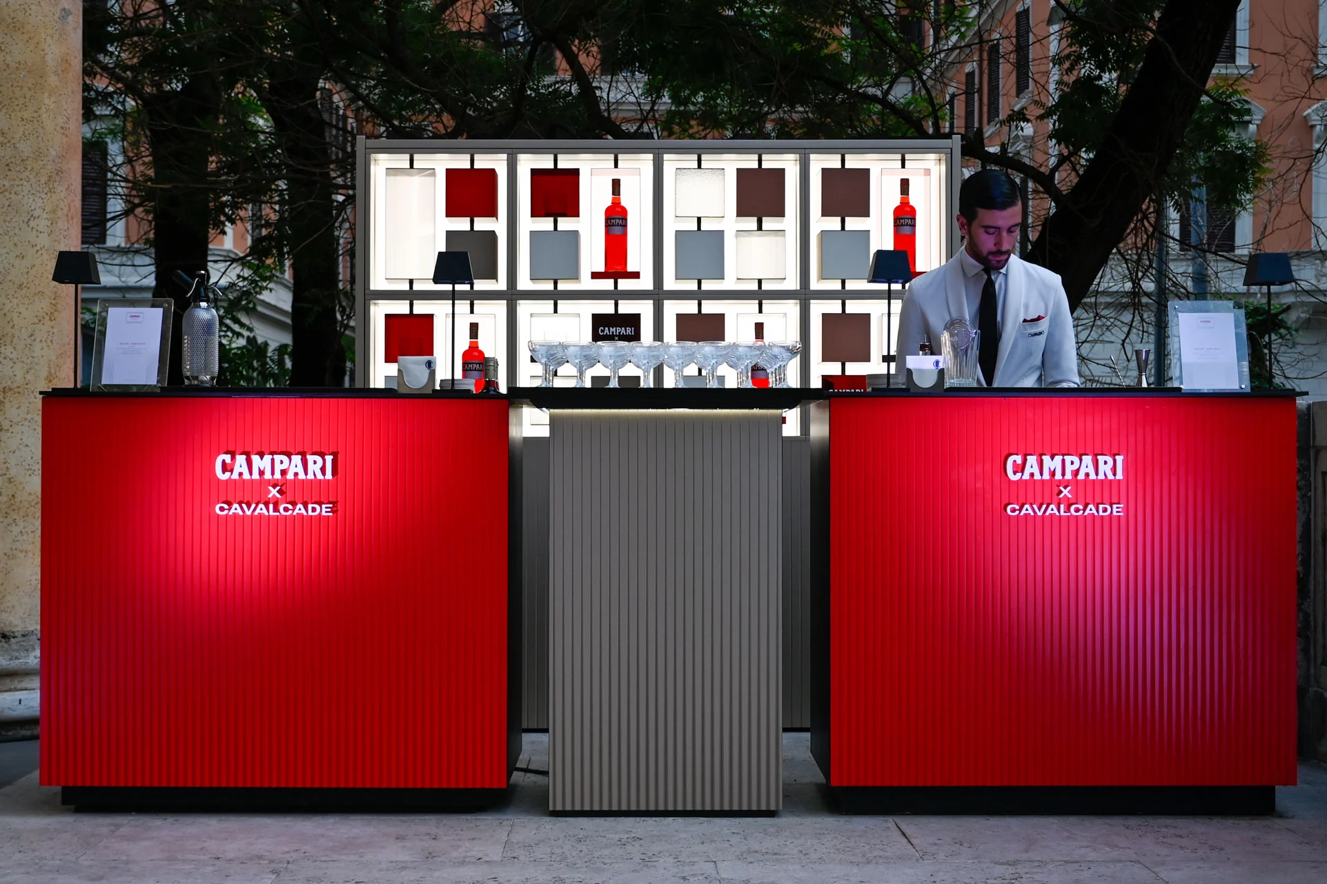 Due banconi bar rossi Campari x Cavalcade davanti a una scaffalatura retroilluminata, con un barman – Campari Cavalcade, Roma, Italia, 2023 – progettazione di eventi