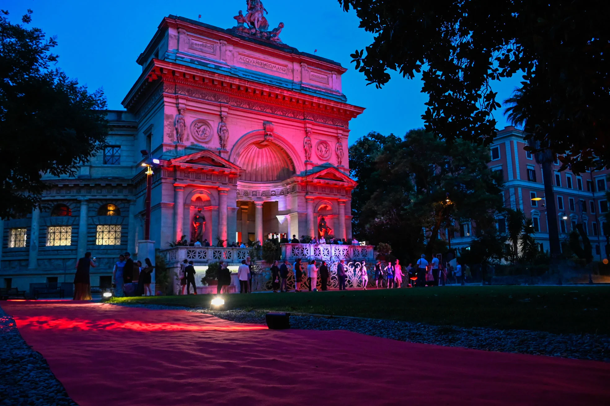 Edificio storico ad archi illuminato di rosa al tramonto, ospiti riuniti in fondo a un lungo tappeto rosso – Campari Cavalcade, Roma, Italia, 2023 – progettazione di eventi