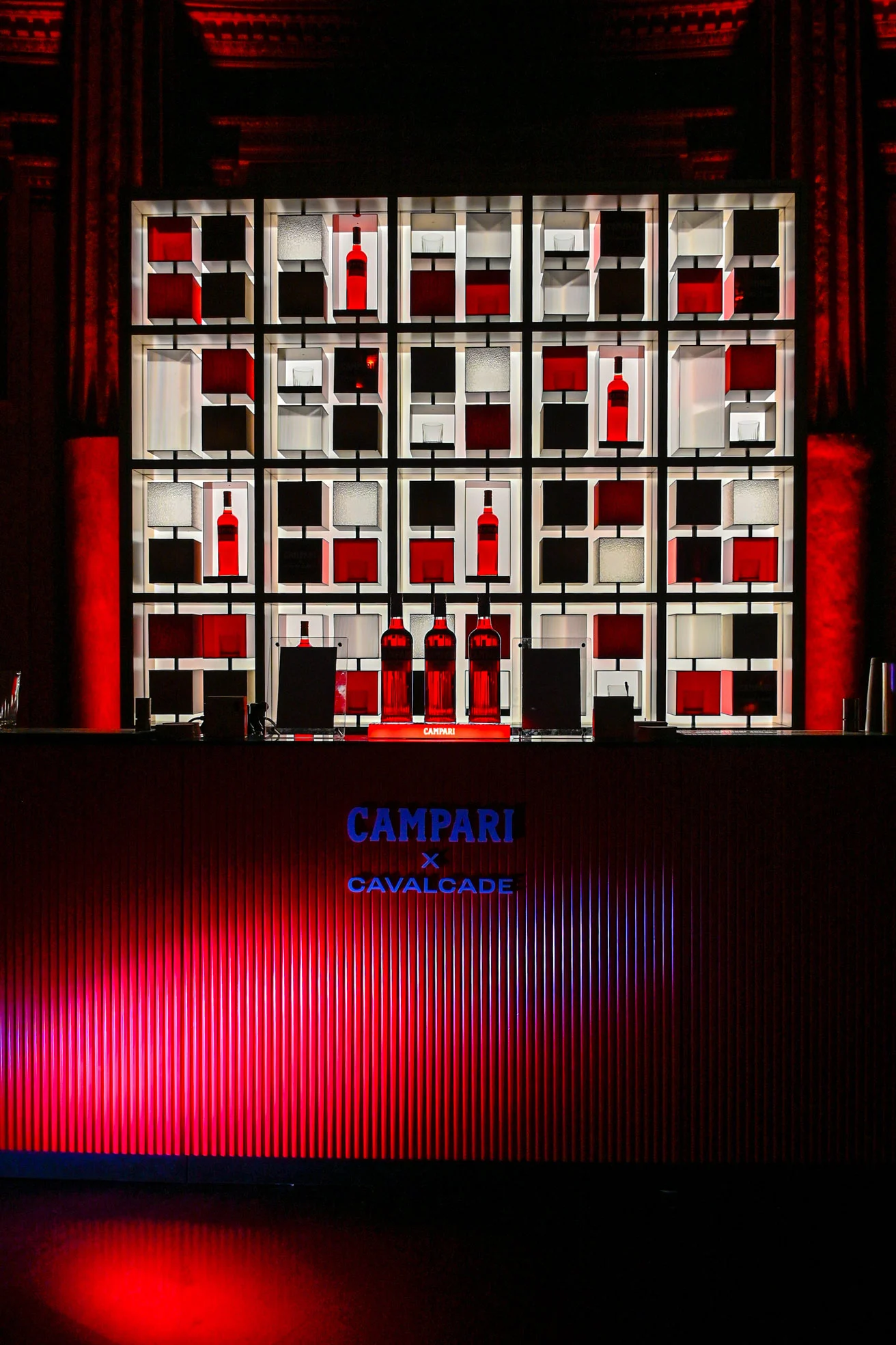 Scaffalatura a griglia retroilluminata con bottiglie Campari dietro un bancone bar Campari x Cavalcade illuminato di rosso – Campari Cavalcade, Roma, Italia, 2023 – progettazione di eventi