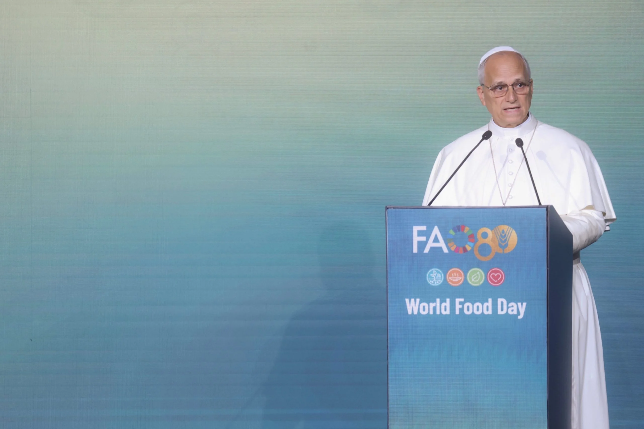 Oratore in abiti bianchi al podio FAO80 della Giornata mondiale dell'alimentazione davanti a un fondale verde acqua – FAO MuNe Museo dell'alimentazione e dell'agricoltura, Roma, Italia, 2025 – allestimento espositivo