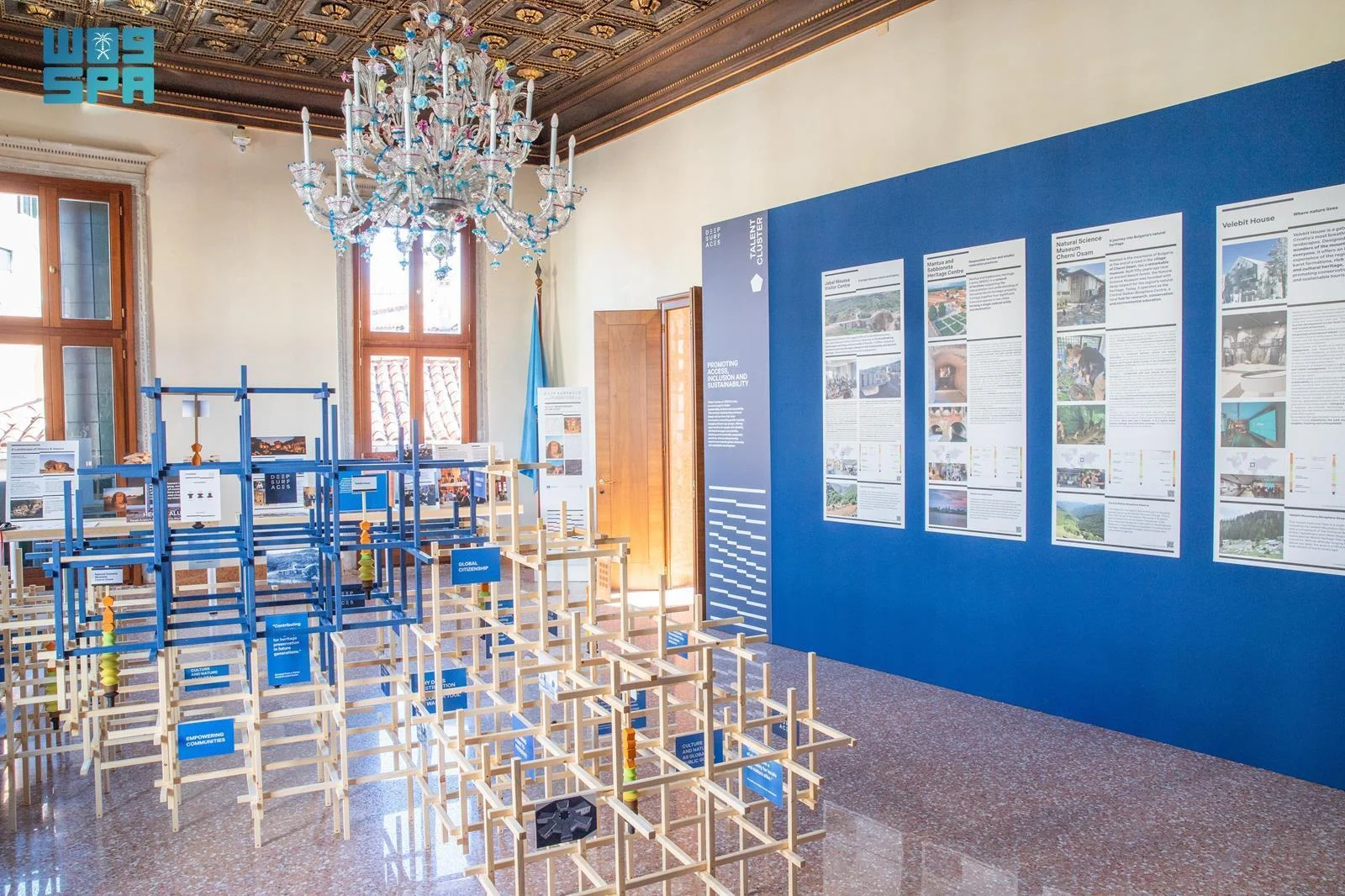 Sala espositiva con strutture a graticcio in legno blu e naturale sotto un lampadario di Murano, pannelli dei progetti su una parete blu – Deep Surfaces a Palazzo Zorzi, Venezia, Italia, 2025 – allestimento espositivo