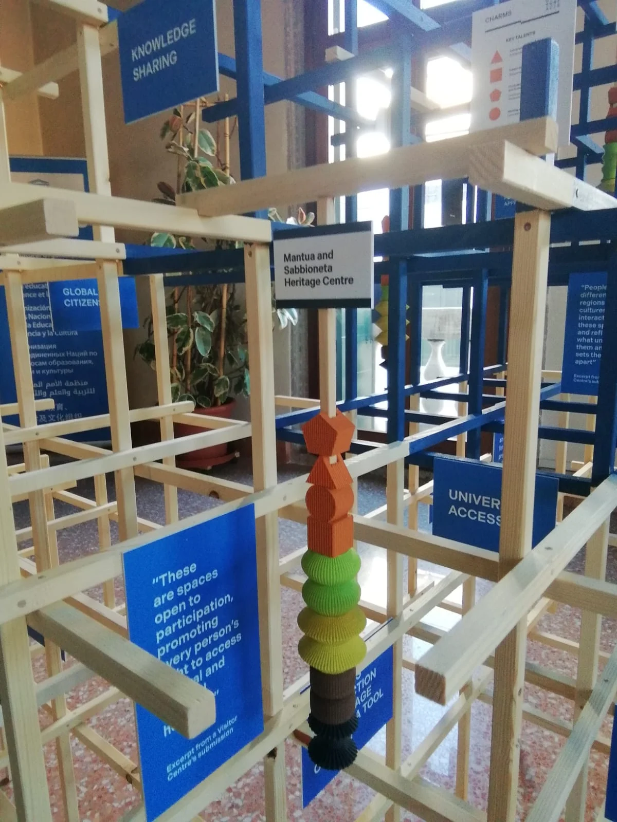 Dettaglio all'interno del graticcio in legno: una pila di pezzi colorati stampati in 3D tra didascalie blu ed etichette dei progetti – Deep Surfaces a Palazzo Zorzi, Venezia, Italia, 2025 – allestimento espositivo