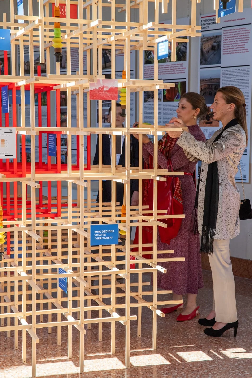 Visitatori che inseriscono un pezzo nell'alta installazione a graticcio in legno, luce del sole sul pavimento di marmo – Deep Surfaces a Palazzo Zorzi, Venezia, Italia, 2025 – allestimento espositivo
