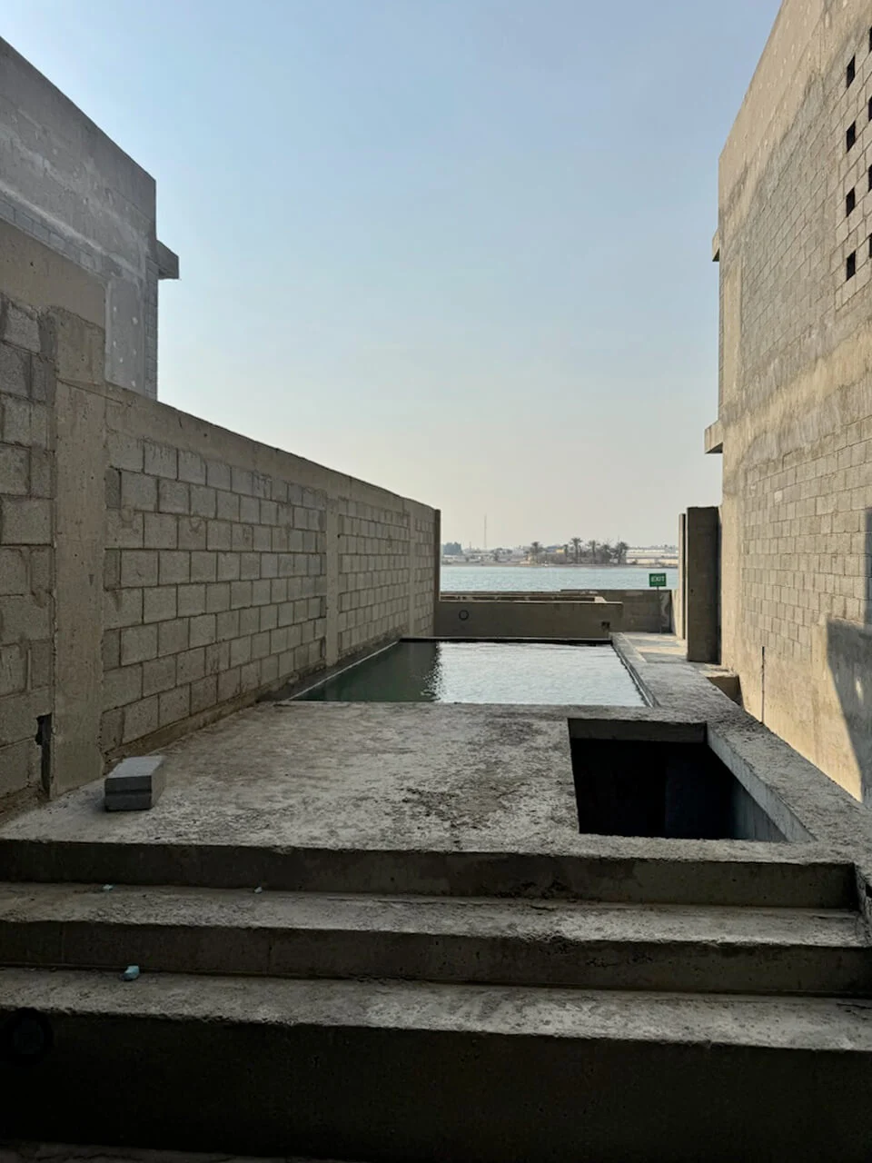 Gradini in cemento che portano a una piscina incompiuta tra muri in blocchi, con il mare oltre – Radic Residences, Jeddah, Arabia Saudita, 2025 – architettura, in costruzione