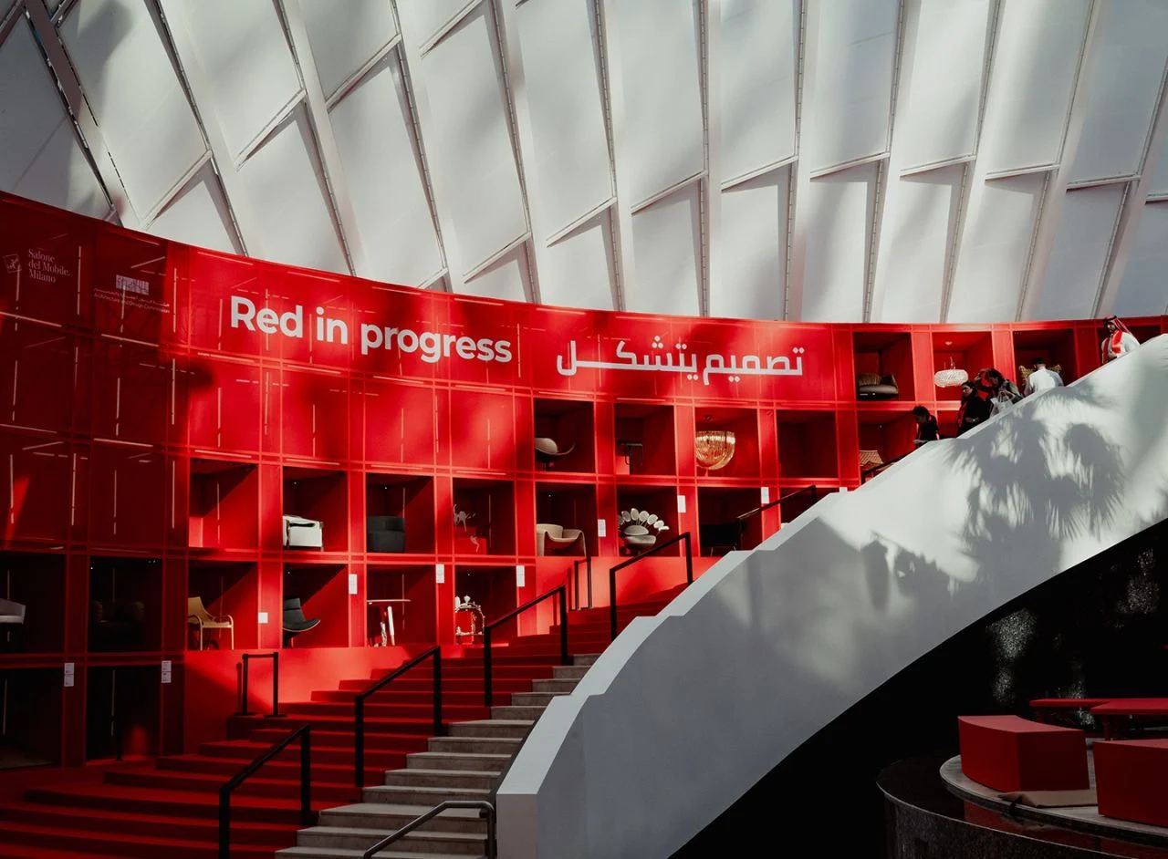 Parete di scaffali rossa con la scritta Red in progress e arredi esposti, accanto a una scala rossa sotto una copertura bianca – Red in Progress: Salone del Mobile Meets Riyadh, Arabia Saudita, 2025 – allestimento espositivo